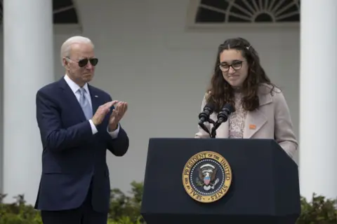 Bloomberg via Getty Images US President Joe Biden applauds as Mia Tretta, a victim of gun violence, speaks during an event in the Rose Garden of the White House in April 2022. BIden is wearing a navy suit and blue tie and Mia is wearing a pink dress and coat and stnding in from of a podium