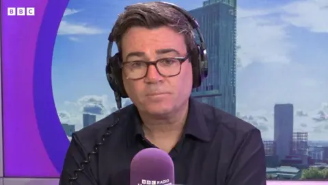Andy Burnham, a man with dark hair in a black shirt, talks into a purple microphone on BBC Radio Manchester. 