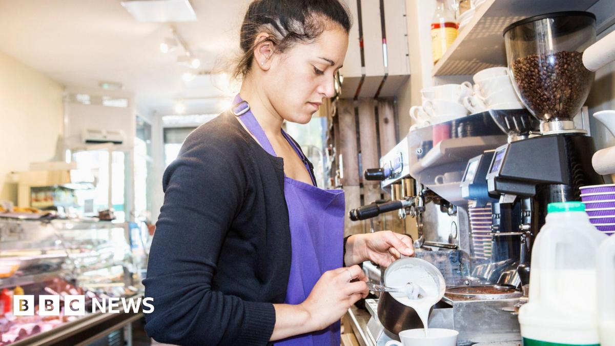 A woman wearing a purple apron works in a cafe. She is pouring milk into a mug next to a coffee machine