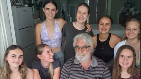 Family photo An older man with grey hair and beard, wearing a black and grey striped t shirt, black shorts and glasses. He is surrounded by seven young girls wearing summery clothes. They are all smiling and looking at the camera.
