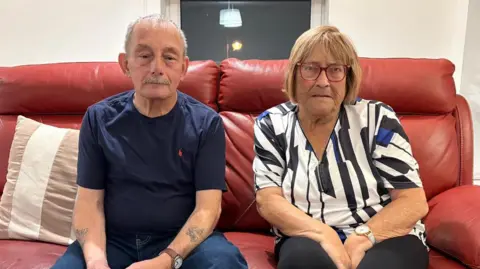 BBC An elderly couple sit on a red leather sofa in a living room. The man on the left has cropped grey hair and a grey moustache, and wears a navy blue t-shirt and dark jeans. The woman has blonde bobbed hair, and wears red-rimmed glasses and a patterned blouse.