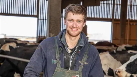 Staffordshire County Council Edward Johnson, a dairy farmer, is seen standing in a cow shed surrounded by cattle. He is wearing a blue jumper and overalls. He is smiling at the camera.