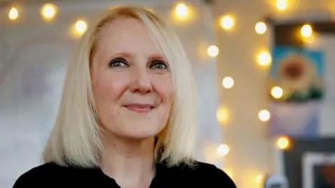 Murdo MacLeod Tressa Burke, who has shoulder length blonde hair, looks off into the distance. There are lights and a picture of a sunflower on the wall behind her.