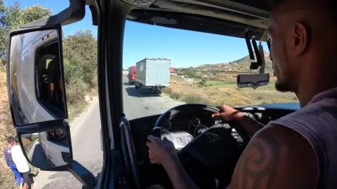A view from the cab of a lorry showing a narrow road and some stationary vehicles.