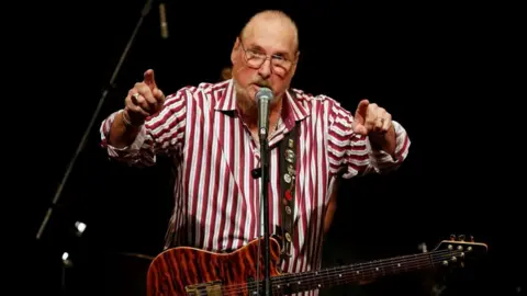 Reuters Steve Cropper, with a brown guitar hanging from his neck, gestures with both hands pointing forward. He wears a red and white striped button-down shirt