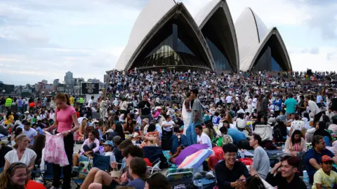 Getty Images Images is filled with people sitting and standing as they gather ahead of the fireworks display. Behind them is the Sydney Opera House and it is daylight still