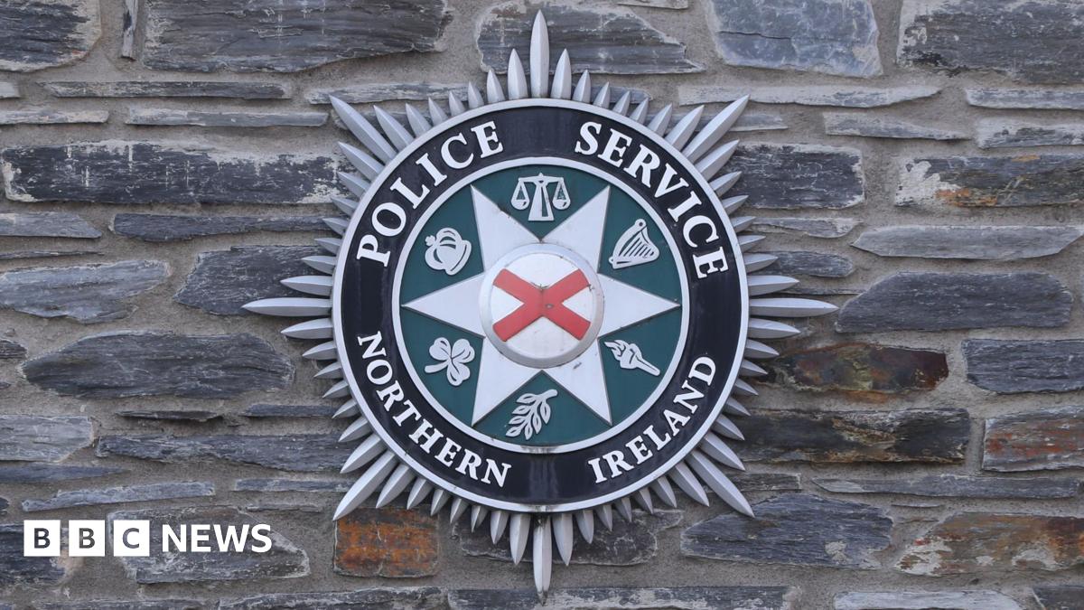 Black, green, and grey circular crest on a grey brick wall, featuring the words Police Service Northern Ireland in white lettering.