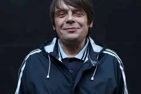 Getty Images Gary "Mani" Mounfield is wearing a dark navy windbreaker jacket with white stripes along the sleeves and white drawstrings hanging from the hood. Underneath the jacket, a collared shirt with a light blue colour and a dark buttoned sweater is visible. The background is solid black, creating a simple and minimalistic setting.
