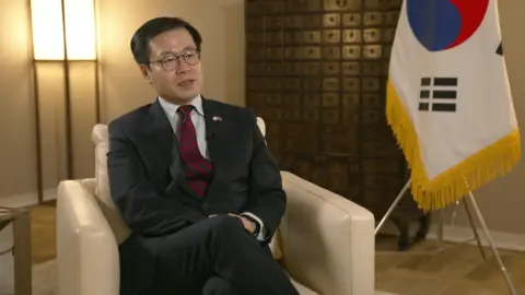 South Korea's trade minister Yeo Han-koo sits in a cream leather chair with his country's flag in the background as he is interviewed by a BBC reporter.