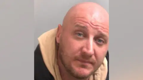 Essex Police Custody picture of Charlie Lancaster. He has a shaved head and stubble and is wearing a beige hoodie and black jacket. He has blue eyes and is looking directly at the camera. 
