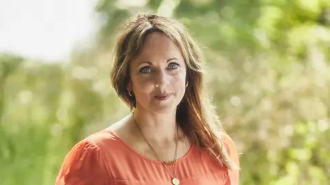 Josh Adam Jones Jenny Evans smiles at the camera. She is wearing a coral pink dress and a necklace. Her blonde hair is pulled to the side and is resting on her shoulder. Behind her is a blurred background of a garden.