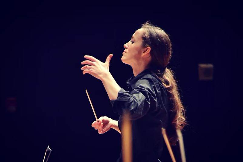 Jessica Cottis in the midst of conducting an orchestra, dressed in all black with her hair tied back in a ponytail.