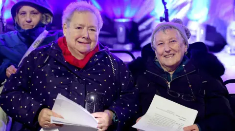 Karbon Homes Two older women in front of a stage which is lit in purple and blue lights. The woman on the left has short, grey hair and is wearing a red scarf with a blue coat which is covered in small hearts. She is smiling and holding sheets of paper. The woman on the right also has short, grey hair but it wearing a grey wooly hat. She is wearing a black coat with a fur hood and is also holding white sheets of paper which look to have lyrics on. She is smiling and her teeth showing. There is a woman standing behind them with blonde hair and her hood up.