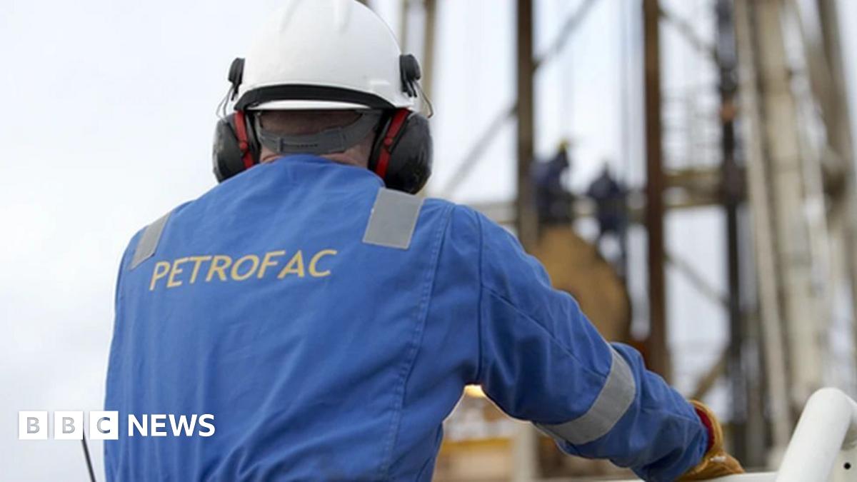 A man in a blue boiler suit and white hard hat looks out over industrial equipment. Petrofac is printed on the back of the boiler suit in capital letters.
