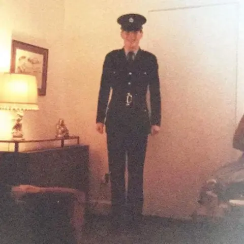 Adrian Hunt A young police cadet standing in full uniform in a warmly-lit living room in the 1980s. The shot is low resolution and taken from far back, but the young man can be seen smiling broadly.