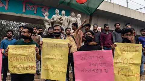 AFP via Getty Images Students with black cloth tied over their faces hold placards during a silent protest to condemn the lynching of Hindu garment worker Dipu Chandra Das near the Raju Memorial Sculpture at Dhaka University in Dhaka on December 21, 2025. Fuelled in part by growing anti-India sentiments in the majority Muslim nation, the violence this week over the killing of student leader Sharif Osman Hadi in Bangladesh saw a Hindu garment worker killed in the central district of Mymensingh on December 18, following allegations of blasphemy. (Photo by Abdul Goni / AFP via Getty Images)