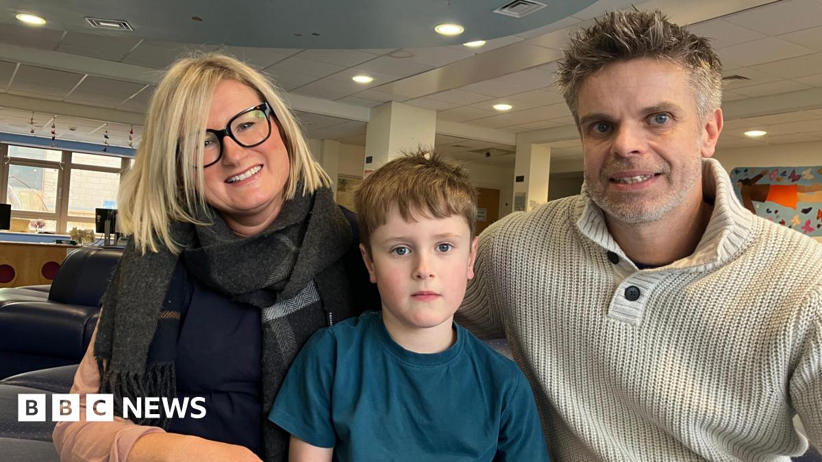 A young boy is sitting between his mum and dad. The boy has short brown hair and is wearing a blue t-shirt.  He is staring at the camera. His mum has dark framed glasses on and blonde short hair.  She is wearing a large scarf and smiling at the camera. The boy's dad is sitting on his left and is wearing a cream knitted jumper, he has grey spikey hair.