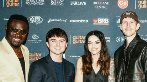 APB PR Four people - three men and one woman - on he red carpet of an awards event. All are dressed up for the event, in suits, back leather jackets or dark tops.