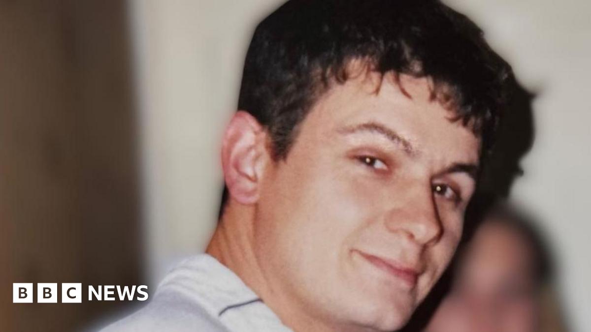 A man with short dark hair turns to the camera. He is wearing a white shirt.