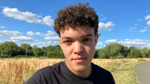 BBC A young man with dark curly brown hair standing by the side of a road. He is wearing a black t-shirt and behind him are yellow crops and blue skies.