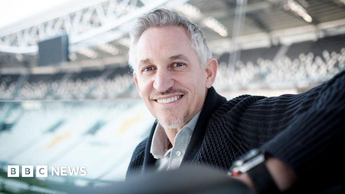 Gary Lineker sits in a football stand smiling, with another stand across the pitch seen behind him out of focus.