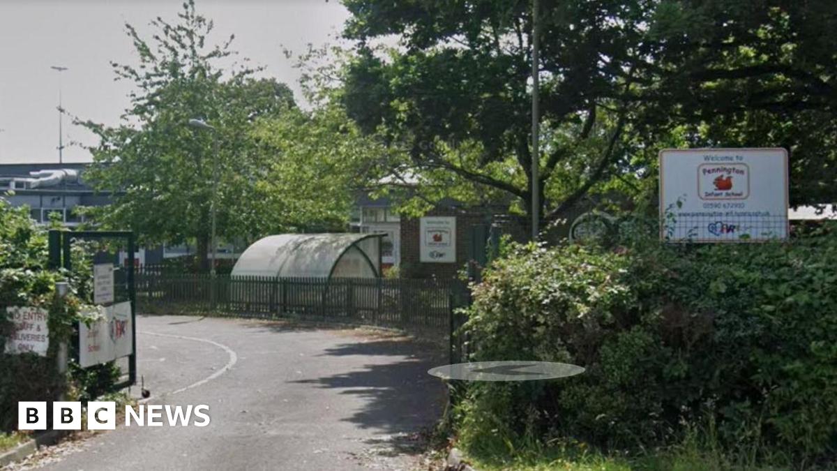 A shot of Pennington Infant School set back among trees and bushes, with open school gates in the foreground, and more trees and bushes to the right of the picture. A plastic bike shelter is in the background.