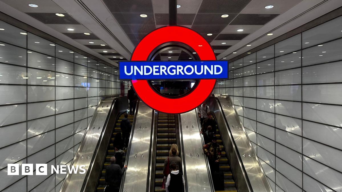 A large Tube roundel hangs above a bank of three escalators as Euston underground station. There are white tiles on the wall and people are visible riding the escalators.