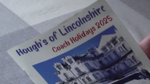 The front cover of a stapled holiday brochure held above a grey and white checked table cloth. The brochure is white and features an image of a large, white hotel beneath a blue sky. Blue text in a comic-sans-style font reads: "Hough's of Lincolnshire". Another line of text, in red, reads: "Coach Holidays 2025".