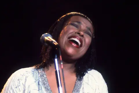 Getty Images Roberta Flack is performing on stage with a microphone on a stand in front of he. She is wearing a dark outfit with a high neckline and has shoulder-length hair styled straight. The background is dark with a soft spotlight illuminating the figure, creating a dramatic concert atmosphere.