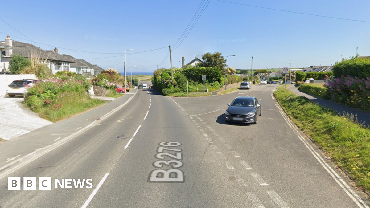 The junction where the B3276 Porth Way meets Well Way. There is a car waiting to drive out of Well Way. There is a row of bungalows on the left of Porth Way. The sea can be seen on the horizon. It is a sunny day with a clear, blue sky.