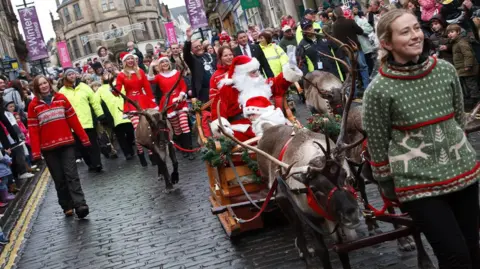Cairngorm Reindeer Herd A woman in a pale green Icelandic-style jumper leads two harnessed reindeer pulling Santa's sleigh, and another two walking behind, during a town centre festive event.