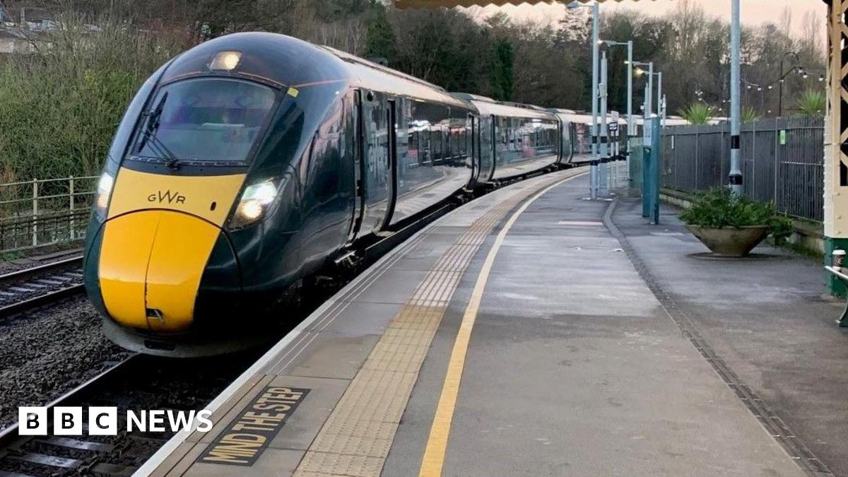 A green GWR train pulls into a railway station. It has its lights on. The platform is empty.