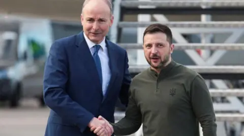 Reuters Volodymyr Zelensky and Micheál Martin shake hands at the steps of a plane at Shannon Airport. Martin is wearing a navy suit with a white shirt and blue tie. Zelensky has a black and grey beard and is wearing a green long sleeve top with a Ukrainian symbol.