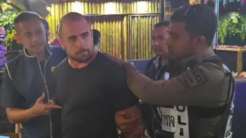 Essex Police Uniformed Royal Thai Police officers arrest Ben Ransome in a restaurant, which has bamboo on the walls. He is wearing a black T-shirt and is biting his lip.