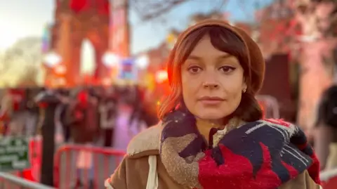 Eve Massie Bishop, One Kind head of campaigns, looks serious at a crowded Christmas event, wearing a brown beret and a colourful scarf. She is a brown-haired woman.
