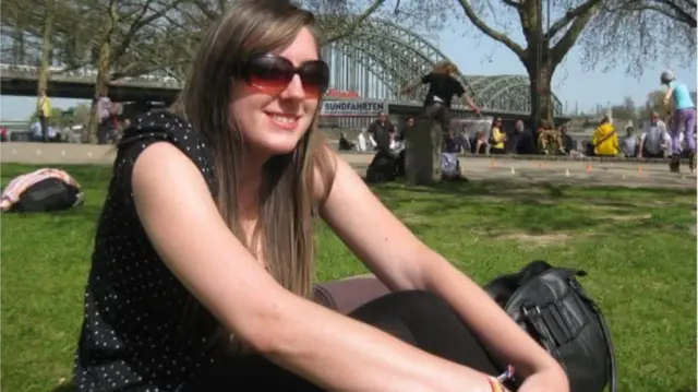 Kellie Snowden wearing a black, polka dot top and sunglasses, sitting in a park with a bridge behind her.