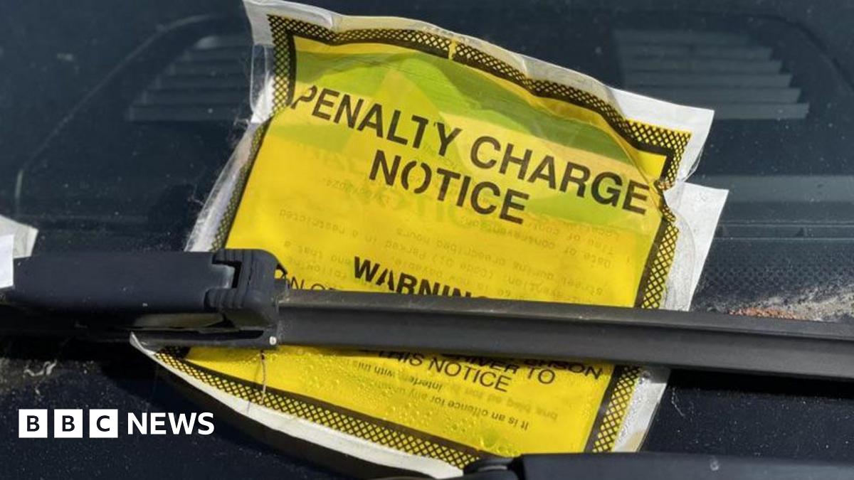 A close-up of a yellow notice saying penalty charge notice and warning. It is tucked under a windscreen wiper and leaning against a windscreen.