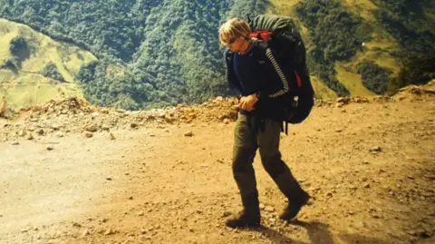 Karl Bushby A man with long blond hair and stubble walks along a mountain pass with dense forest below. He is carrying a large rucksack with a green cover over it.