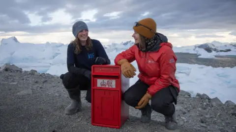 BAS/Jake Martin Kirsten Shaw and Aurelia Reichardt are kneeling beside a red postbox on an outcrop of rocks and shingle. They are both wearing woolly hats and jackets and there is snow on the landscape in the background.