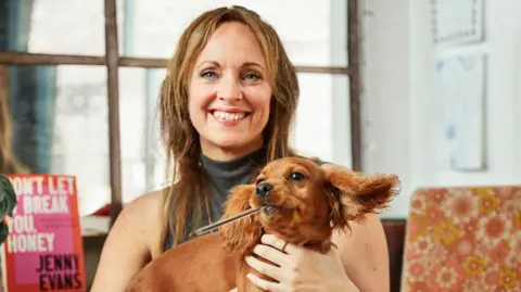 Josh Adam Jones Jenny Evans smiles at the camera and she is holding a brown puppy which has a pen in it's mouth. Behind Jenny is a book that says 'Don't let it break you, honey' by Jenny Evans. 