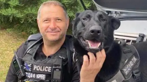 West Midlands Police A man with short grey hair and wearing a black police officer's uniform, smiles as he sits on the back of a car. A black-coloured dog is next to him. The man is scratching the dog under his chin.