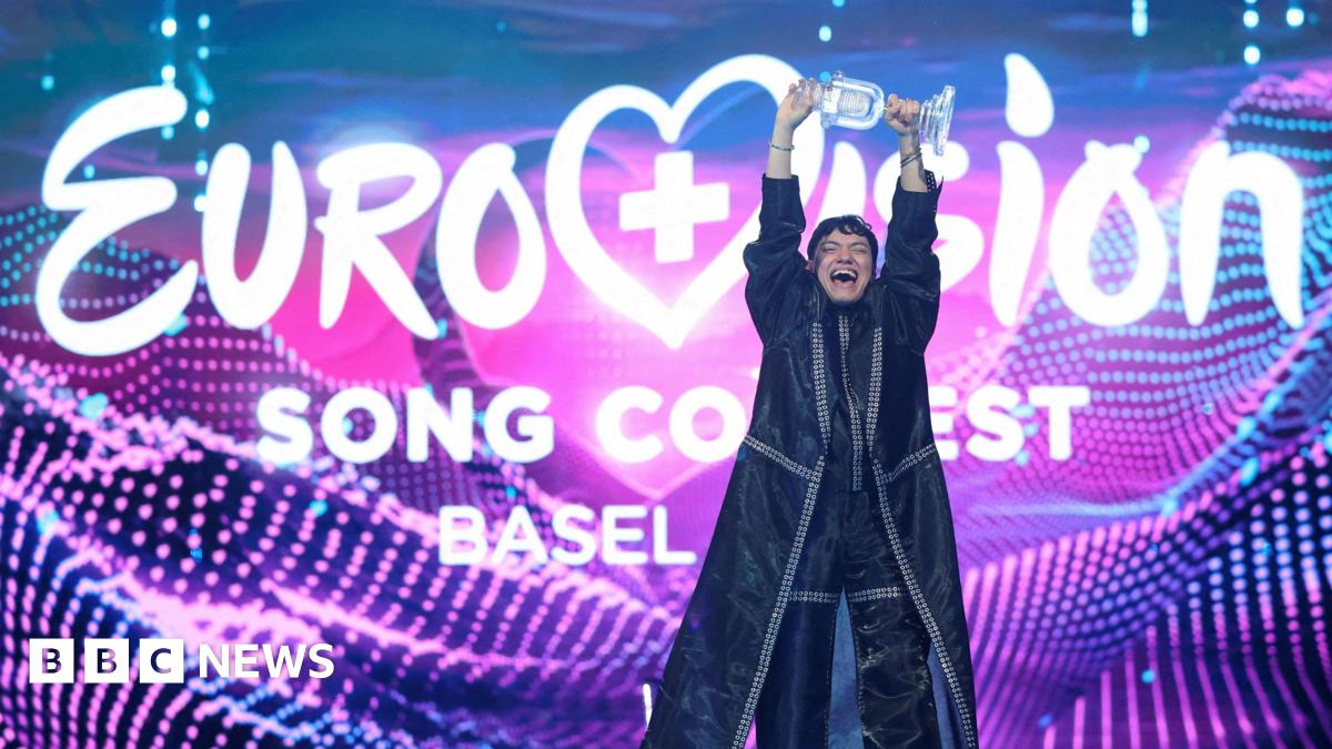JJ holding aloft the trophy in celebration at Eurovision 2025 in front of a giant colourful Eurovision Song Contest branded backdrop