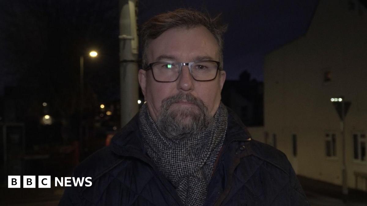 A man with a beard, black glasses, and short dark hair. He is wearing a coat and a grey scarf.