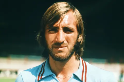 Getty Images Billy Bonds is wearing a light blue shirt with red and white stripes on the collar, standing outdoors in front of a stadium.