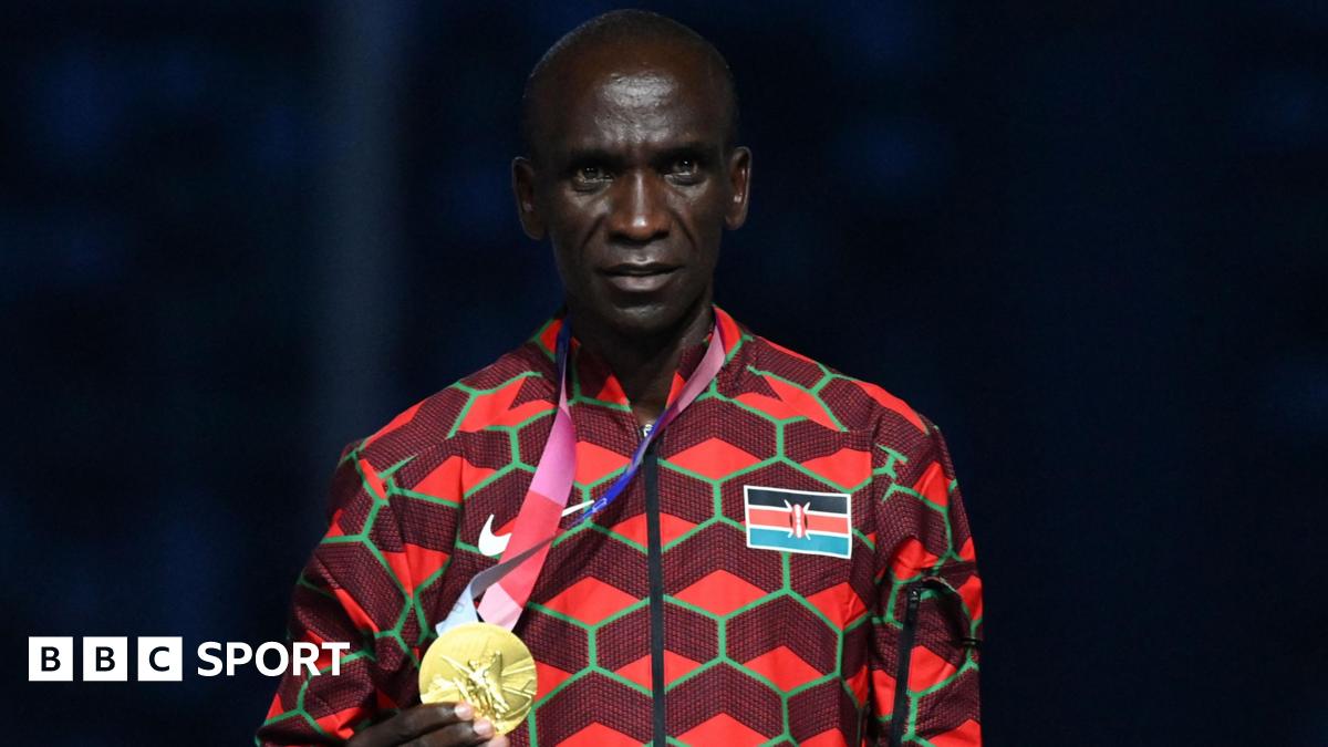 Eliud Kipchoge, visible from stomach up and wearing an official Kenya tracksuit which has the flag on its left breast and is a mainly red design with green hexagonal shapes, poses with his gold medal which he is holding in his right hand and wearing on a ribbon around his neck during the victory ceremony of the men's marathon event during the Tokyo 2020 Olympic Games