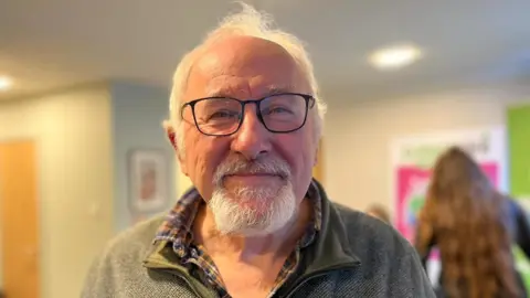 Alasdair Wylie, a white-haired man with a goatee beard, wears a light green fleece and checked shirt and smiles while standing in the Dunkeld cafe.