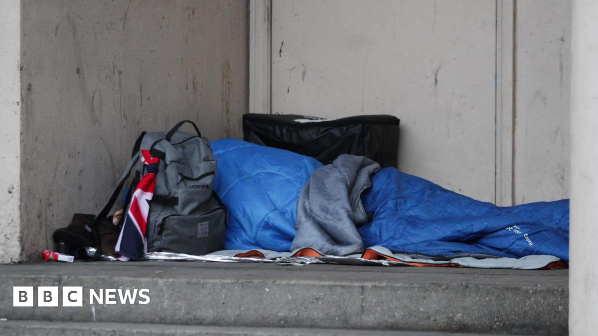 A homeless person sleeping rough in a doorway. Their face and body is covered by a light blue sleeping bag and a grey rucksack blocks view of the person's head.