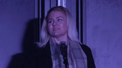 Victims Minister Alex Davies-Jones with long blond hair in a headband wearing a patterned scarf and black coat in a service inside Heaton Park Synagogue