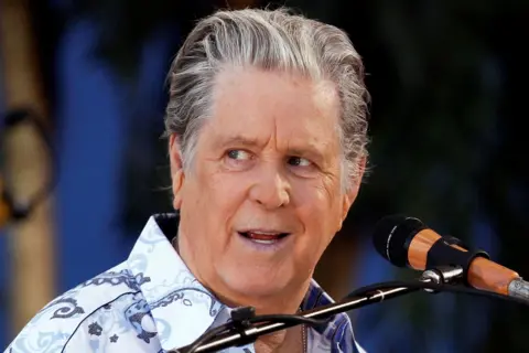 Getty Images Brian Wilson is positioned behind a microphone stand, wearing a light-coloured shirt with a bold paisley pattern in shades of blue and gray. The microphone has a black foam cover and a wooden accent near the mount. The background is softly blurred with dark green and blue tones, suggesting an outdoor or stage setting with natural or ambient lighting.
