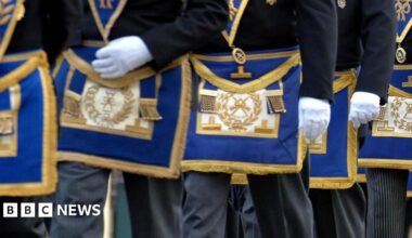 Masons march in a procession. Pictured are men from waist height wearing white gloves and wearing the Freemasons coat of arms embraided with gold thread on a royal blue background